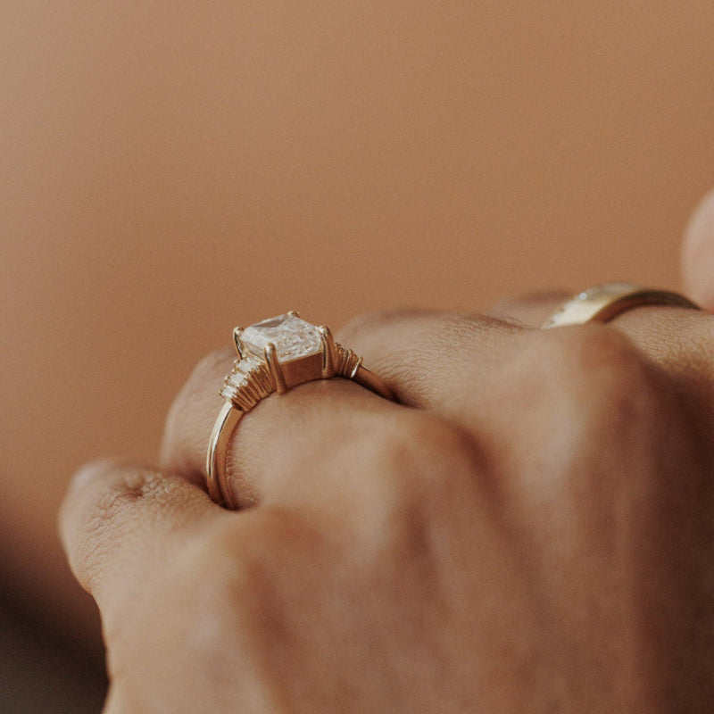The Jolee Emerald Cut Ring in 14k yellow gold worn on the ring finger, shown from a side angle with a wide gold band visible on the middle finger.
