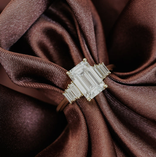 The Jolee Emerald Cut Ring in 14k yellow gold, featuring an emerald cut center stone set in four prongs with baguette natural diamond side stones, displayed on draped brown satin fabric.