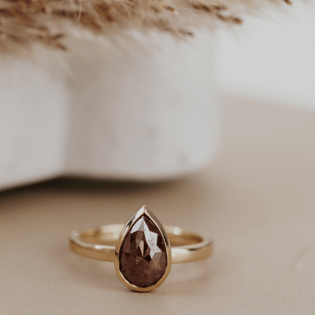 A gold ring with a faceted pear-shaped red rustic diamond, resting on a beige surface with a soft-focus background.