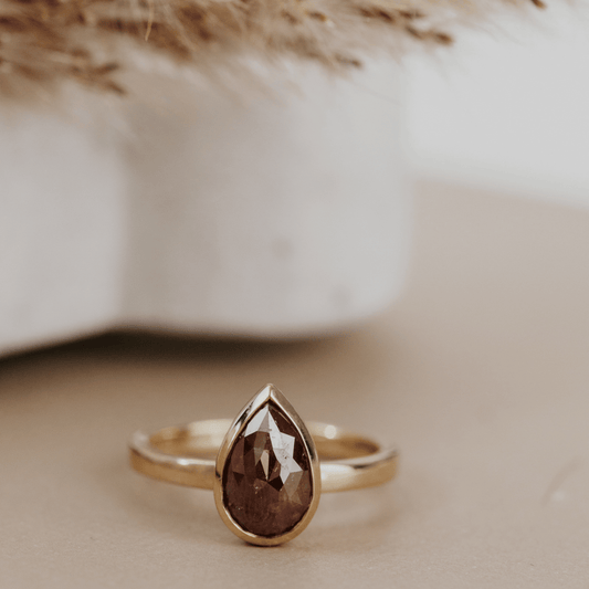 A gold ring with a faceted pear-shaped red rustic diamond, resting on a beige surface with a soft-focus background.