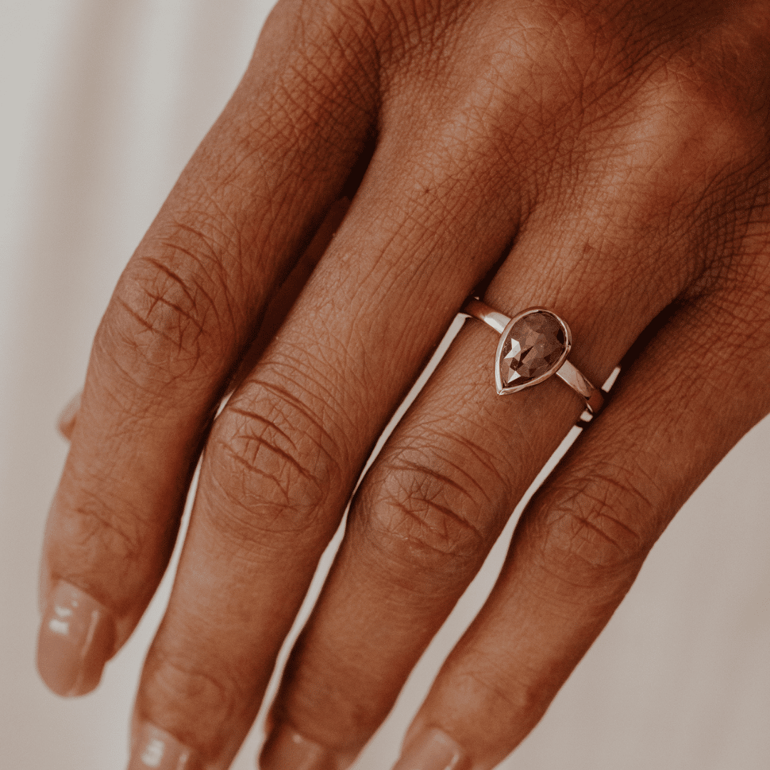 A close-up of a hand wearing a delicate yellow gold ring with a pear-shaped red rustic diamond on the ring finger, set against a soft, warm beige background.