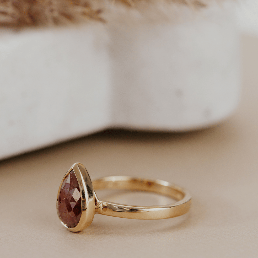 A gold ring with a pear-shaped faceted brown rustic diamond, resting on a beige surface with a soft-focus white ceramic object in the background.