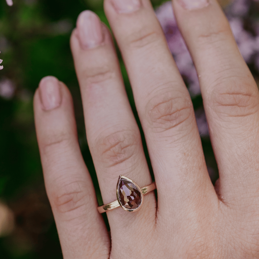 A gold ring with a pear-shaped brown rustic diamond worn on a person's ring finger, shown in a close-up shot against a blurred green and purple background.