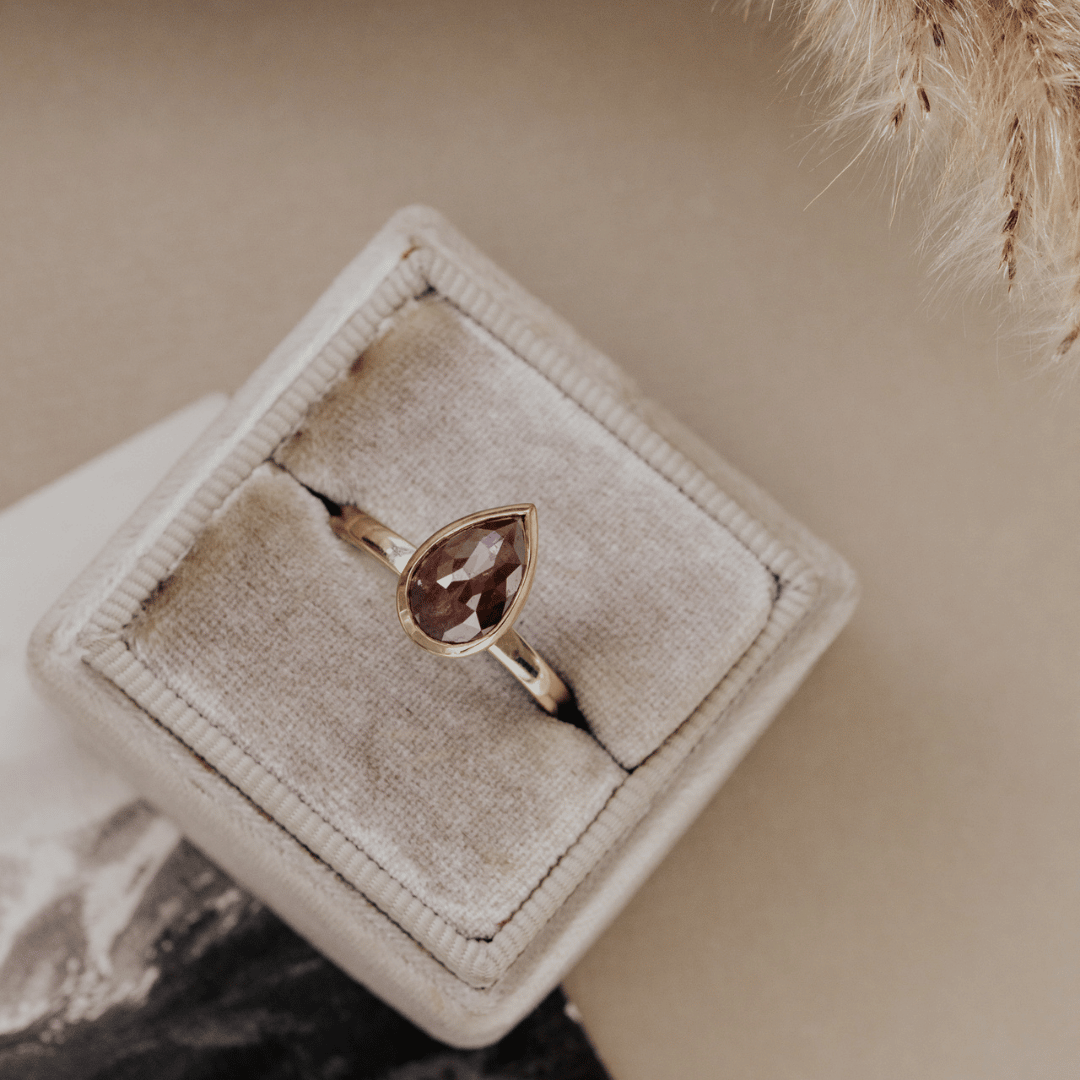 A gold ring with a pear-shaped brown rustic diamond displayed in a beige velvet ring box on a neutral beige background.