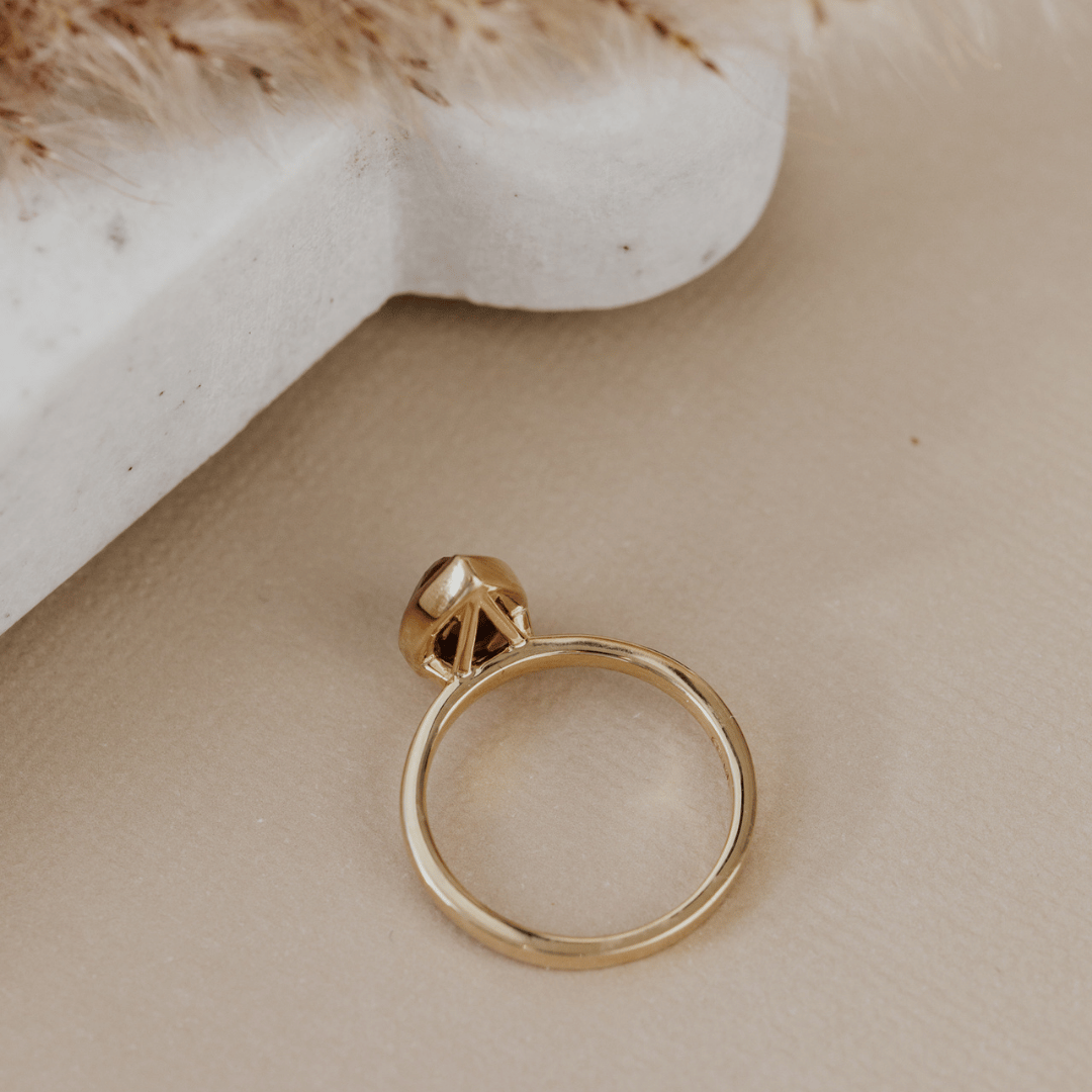 A gold ring with a small geometric triangular accent on the band, photographed on a warm beige surface with a white textured prop in the background.