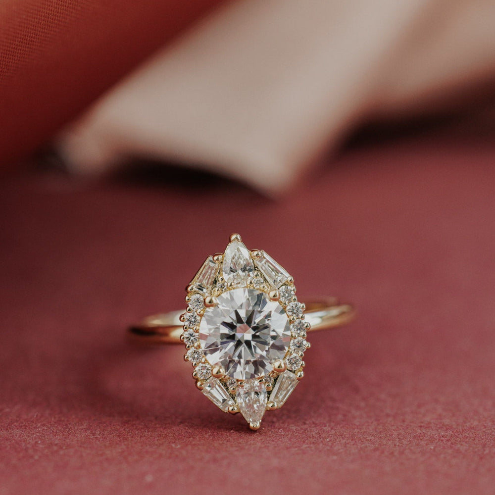 The Lori Round Ring in 14k yellow gold, featuring a round cut center stone in a prong setting surrounded by an ornate halo of pear, baguette, and round natural diamonds, shown from a front-facing angle on a deep red surface.