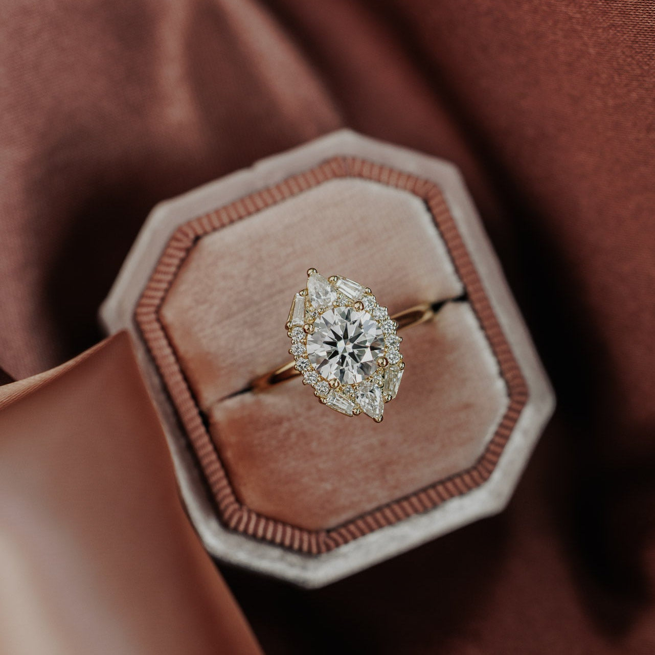 The Lori Round Ring in 14k yellow gold, featuring a round cut center stone in a prong setting surrounded by an ornate halo of pear, baguette, and round natural diamonds, displayed in an octagonal velvet ring box.
