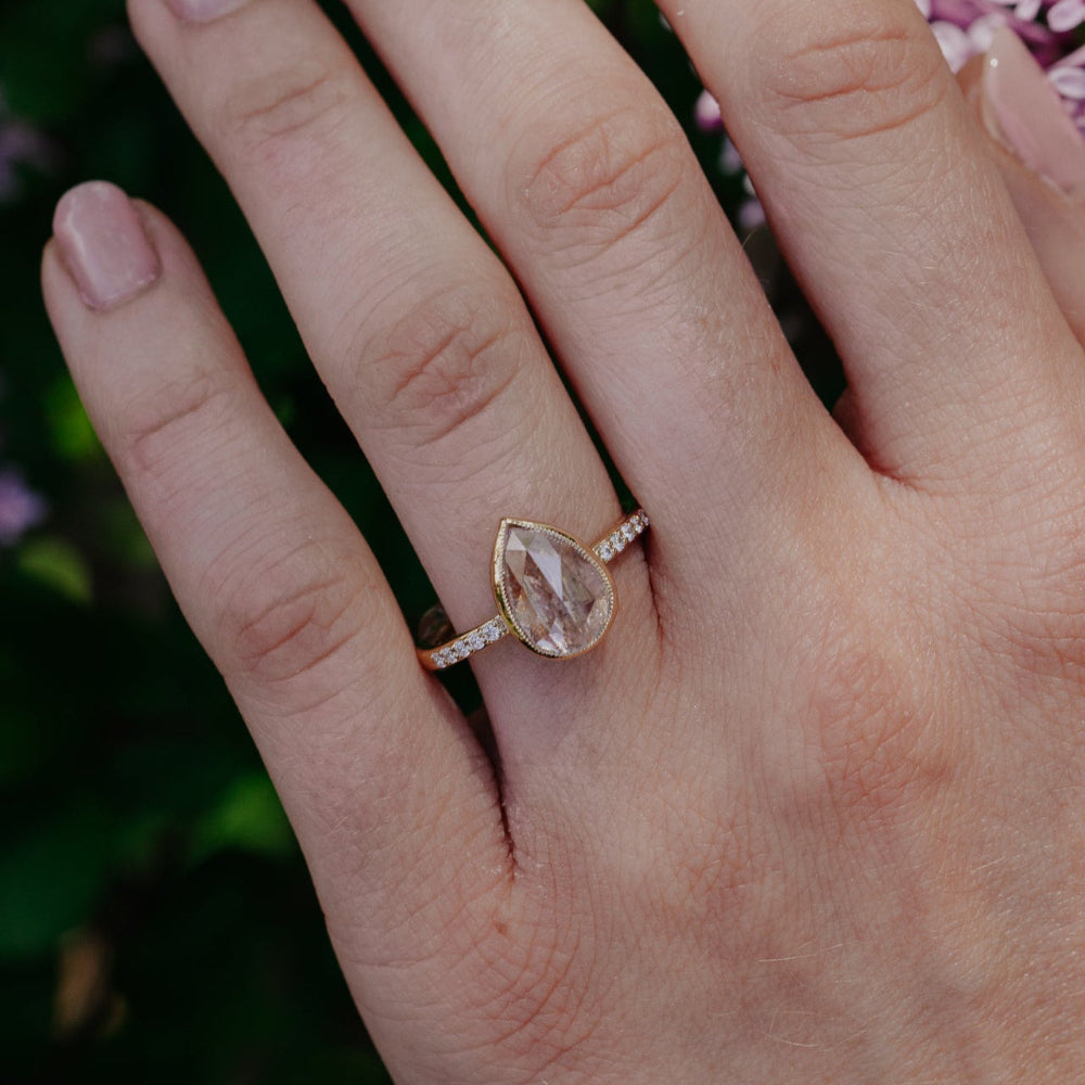 The Lyell 3.24 ct Pear Clear and Peach Rustic Diamond Ring in 14k yellow gold featuring a pear-shaped rose cut rustic diamond in a milgrain bezel with pave diamond band, worn on the ring finger.
