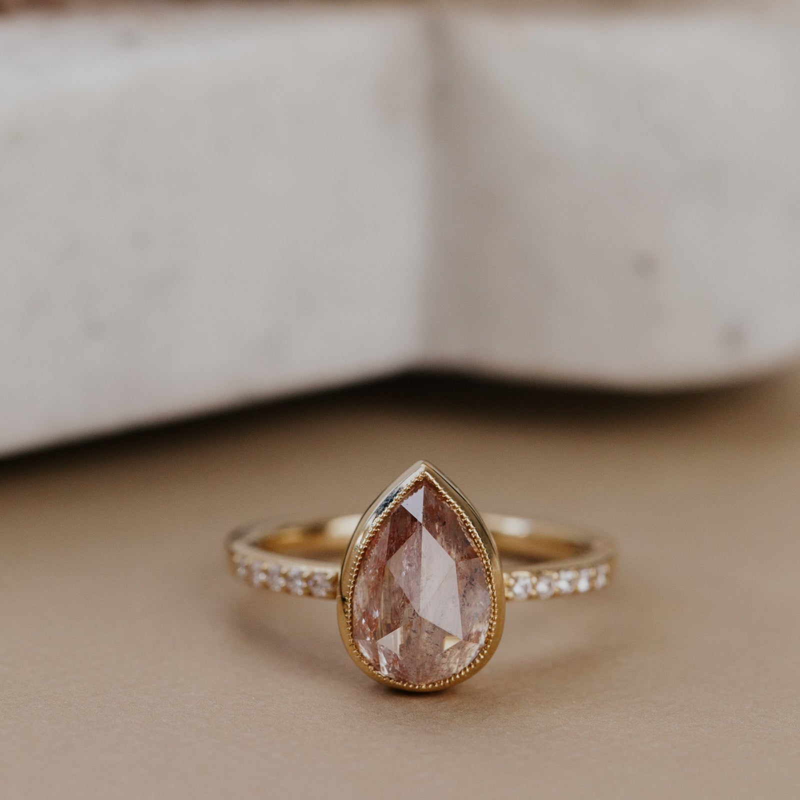 The Lyell 3.24 ct Pear Clear and Peach Rustic Diamond Ring in 14k yellow gold featuring a pear-shaped rose cut rustic diamond in a milgrain bezel with pave diamond band, displayed on a cream surface. This one-of-a-kind 14k yellow gold ring features a 3.24 ct pear-shaped rose cut rustic natural diamond set in a milgrain bezel with ~.09 cttw pave natural diamonds on the band.
