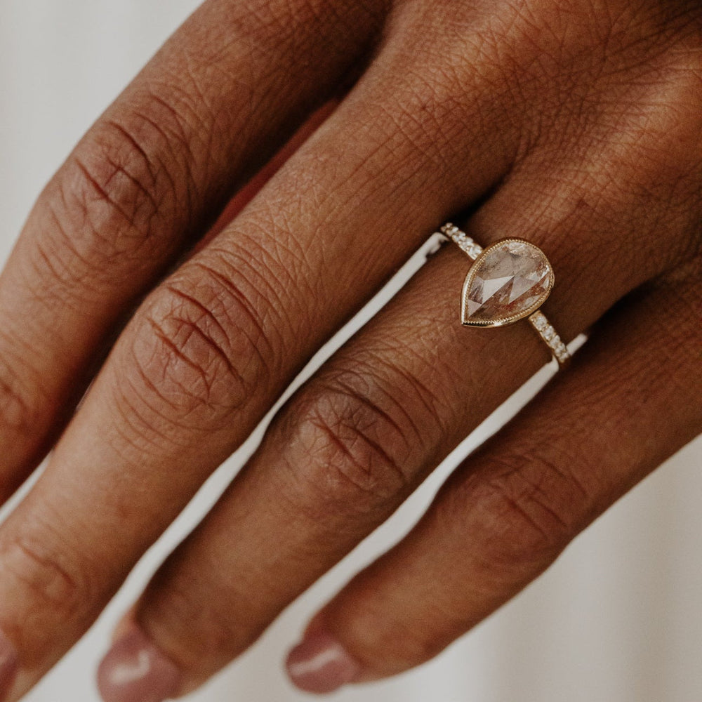 The Lyell 3.24 ct Pear Clear and Peach Rustic Diamond Ring in 14k yellow gold featuring a pear-shaped rose cut rustic diamond in a milgrain bezel with pave diamond band, worn on the ring finger.