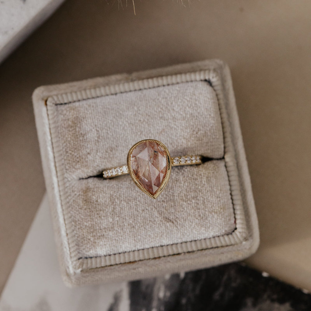 The Lyell 3.24 ct Pear Clear and Peach Rustic Diamond Ring in 14k yellow gold featuring a pear-shaped rose cut rustic diamond in a milgrain bezel with pave diamond band, displayed in a tan velvet ring box. This one-of-a-kind 14k yellow gold ring features a 3.24 ct pear-shaped rose cut rustic natural diamond set in a milgrain bezel with ~.09 cttw pave natural diamonds on the band.

