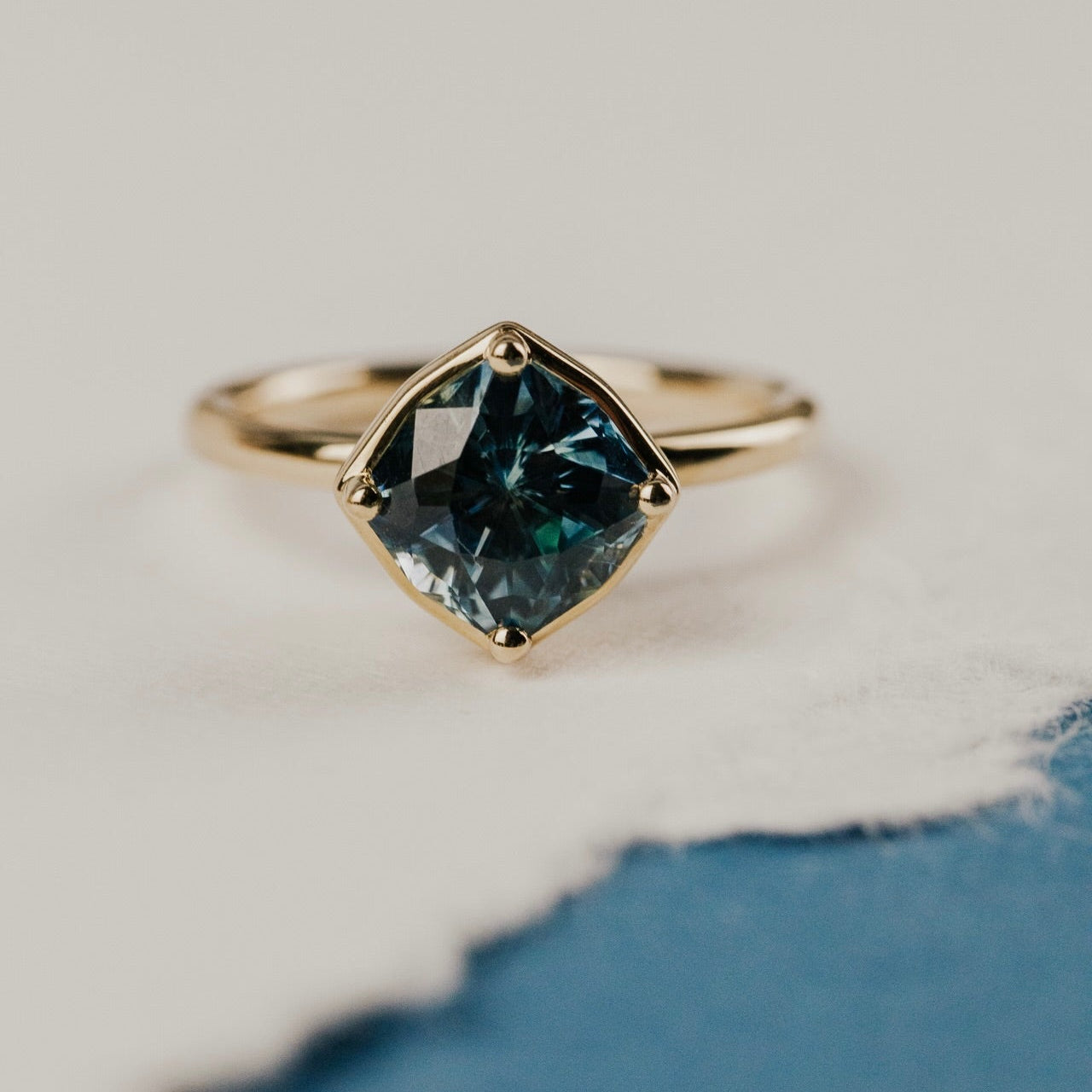 The Maren 2.55 ct Cushion Montana Sapphire Ring in 14k yellow gold featuring a cushion cut Montana sapphire center stone set in four prongs with a high polish gold halo, displayed on cream and blue paper. This 14k yellow gold one-of-a-kind ring features a 2.55 ct fancy cushion cut Montana sapphire set in four prongs with a high polish gold halo.