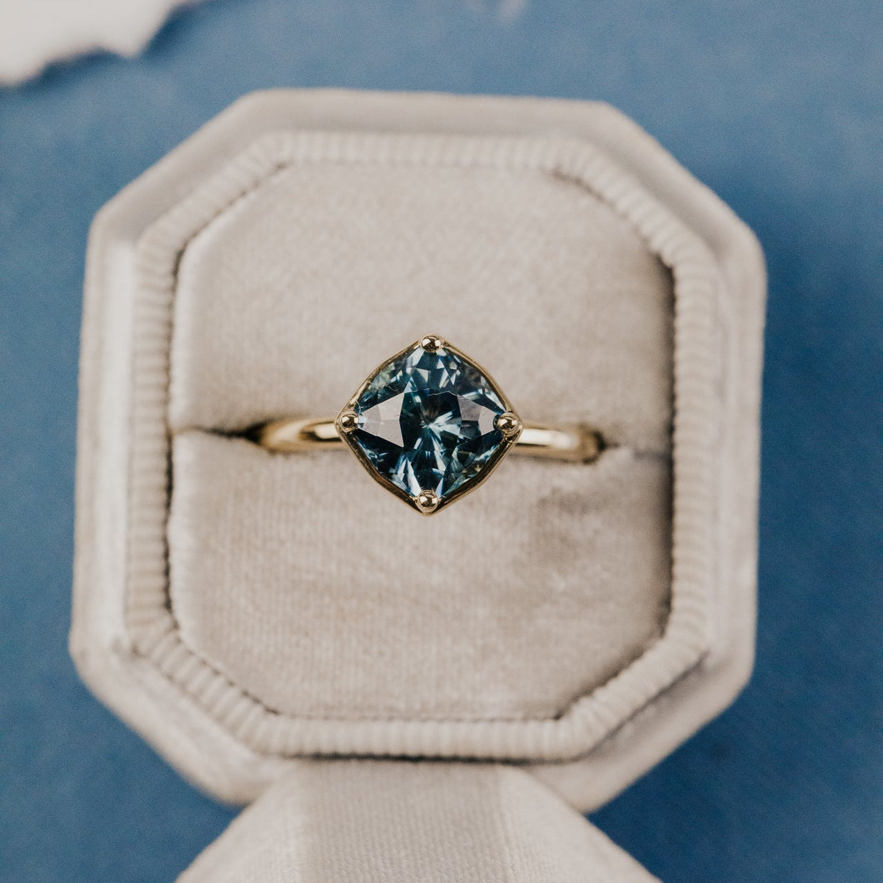 The Maren 2.55 ct Cushion Montana Sapphire Ring in 14k yellow gold featuring a cushion cut Montana sapphire center stone set in four prongs with a high polish gold halo, displayed in a cream velvet ring box. This 14k yellow gold one-of-a-kind ring features a 2.55 ct fancy cushion cut Montana sapphire set in four prongs with a high polish gold halo.
