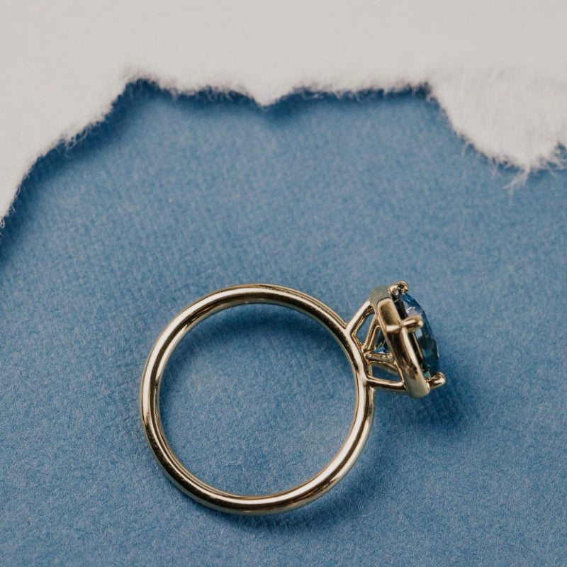 The Maren 2.55 ct Cushion Montana Sapphire Ring in 14k yellow gold featuring a cushion cut Montana sapphire center stone set in four prongs with a high polish gold halo, shown in profile on blue fabric to display the band and setting height.