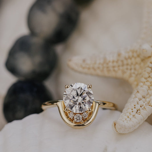 The Palma Round Ring in 14k yellow gold featuring a lab-grown round diamond center stone with round diamond arc accent, displayed on white coral alongside a starfish. The Palma Round Ring is made in 14k gold, featuring a round center stone paired with ~.06 cttw round diamonds set between the center stone and an all-gold arc accent. The entire design is set flush to the finger. This ring is made-to-order based on the selected customizations.