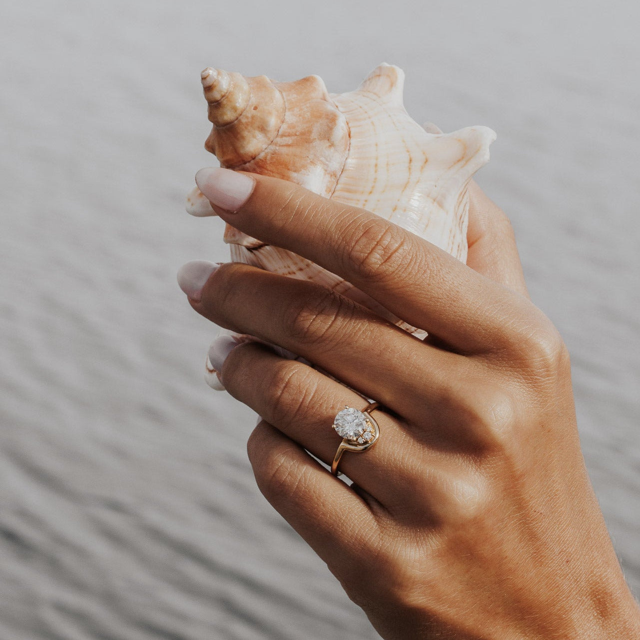 The Palma Round Ring in 14k yellow gold featuring a lab-grown round diamond center stone with round diamond arc accent, worn on the ring finger while holding a seashell.