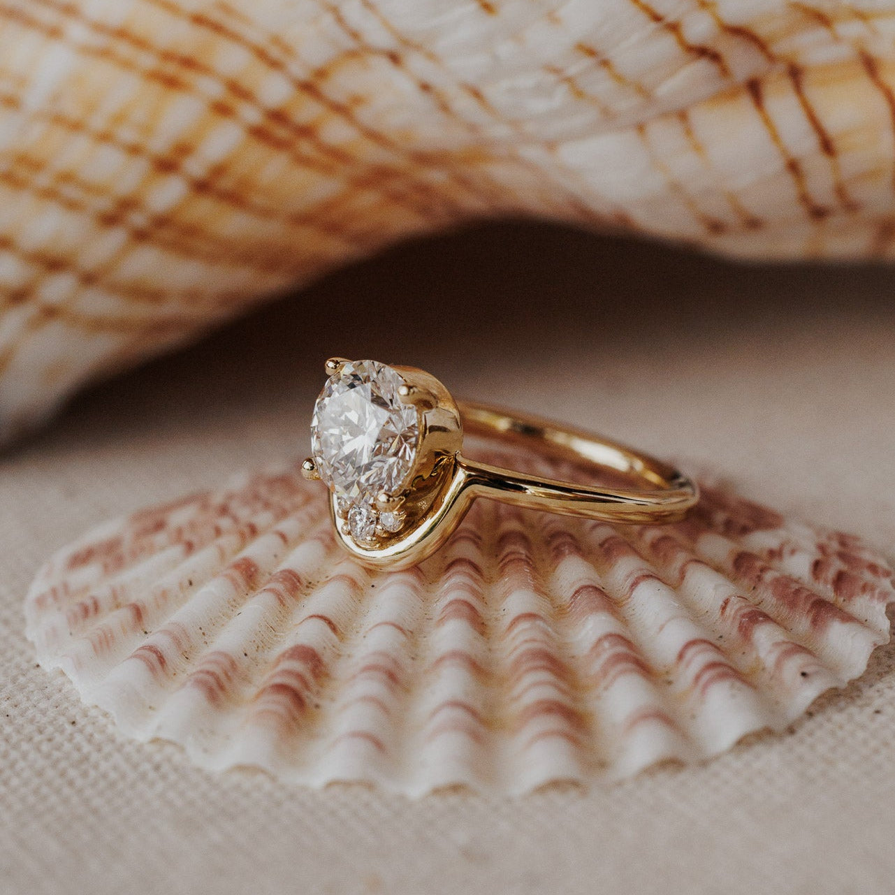 The Palma Round Ring in 14k yellow gold featuring a lab-grown round diamond center stone with round diamond arc accent, displayed on a seashell. The Palma Round Ring is made in 14k gold, featuring a round center stone paired with ~.06 cttw round diamonds set between the center stone and an all-gold arc accent. The entire design is set flush to the finger. This ring is made-to-order based on the selected customizations.
