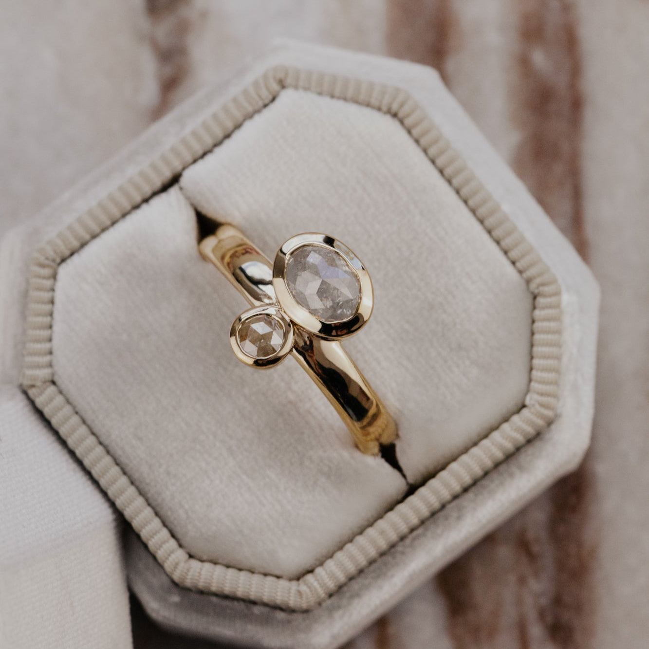 The Reese 1.03 ct Oval and .15 ct Round Rustic Diamond Ring in 14k yellow gold featuring an oval and round rustic diamond set north-south in bezel settings on a wide band, displayed in a cream velvet ring box. This one-of-a-kind 14k yellow gold ring features a 1.03 ct oval rustic natural diamond paired with a 0.15 ct round rustic natural diamond, set north-south in bezel settings on a wide band.
