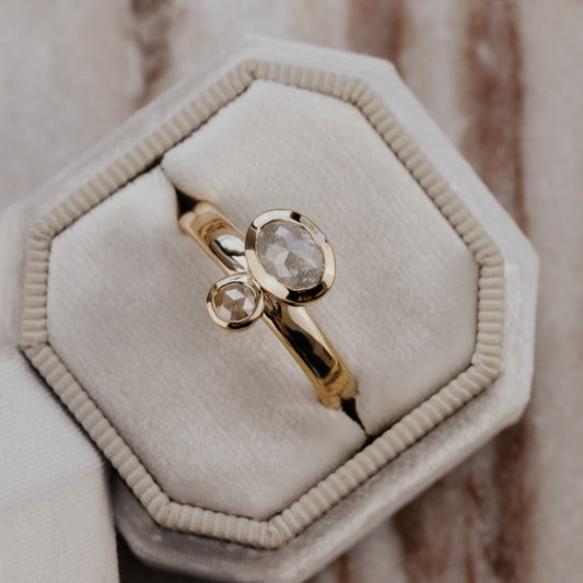 The Reese 1.03 ct Oval and .15 ct Round Rustic Diamond Ring in 14k yellow gold featuring an oval and round rustic diamond set north-south in bezel settings on a wide band, displayed in a cream velvet ring box. This one-of-a-kind 14k yellow gold ring features a 1.03 ct oval rustic natural diamond paired with a 0.15 ct round rustic natural diamond, set north-south in bezel settings on a wide band.
