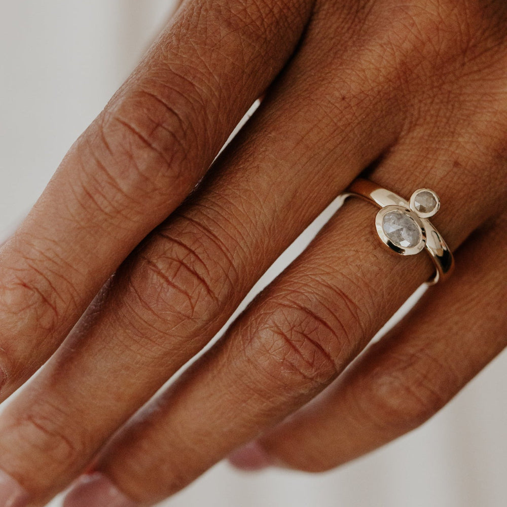 The Reese 1.03 ct Oval and .15 ct Round Rustic Diamond Ring in 14k yellow gold featuring an oval and round rustic diamond set north-south in bezel settings on a wide band, worn on the ring finger.
