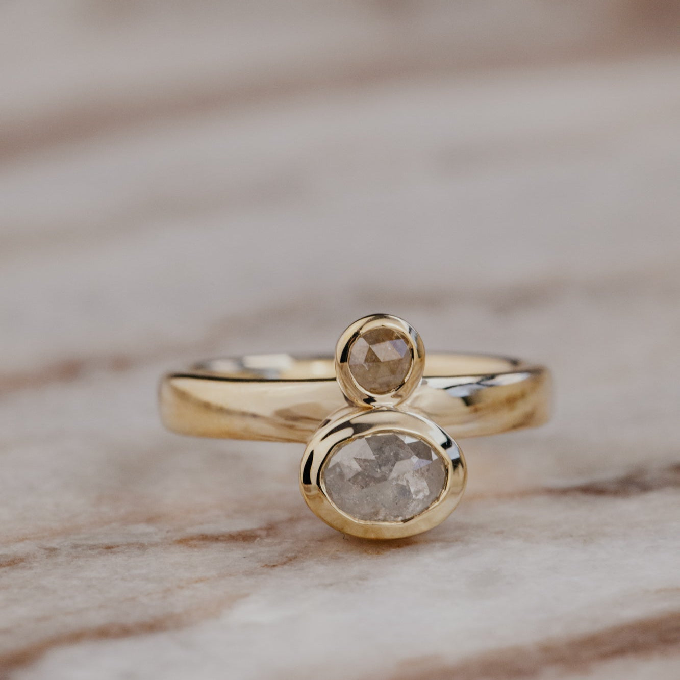 The Reese 1.03 ct Oval and .15 ct Round Rustic Diamond Ring in 14k yellow gold featuring an oval and round rustic diamond set north-south in bezel settings on a wide band, displayed on a marble surface. This one-of-a-kind 14k yellow gold ring features a 1.03 ct oval rustic natural diamond paired with a 0.15 ct round rustic natural diamond, set north-south in bezel settings on a wide band.
