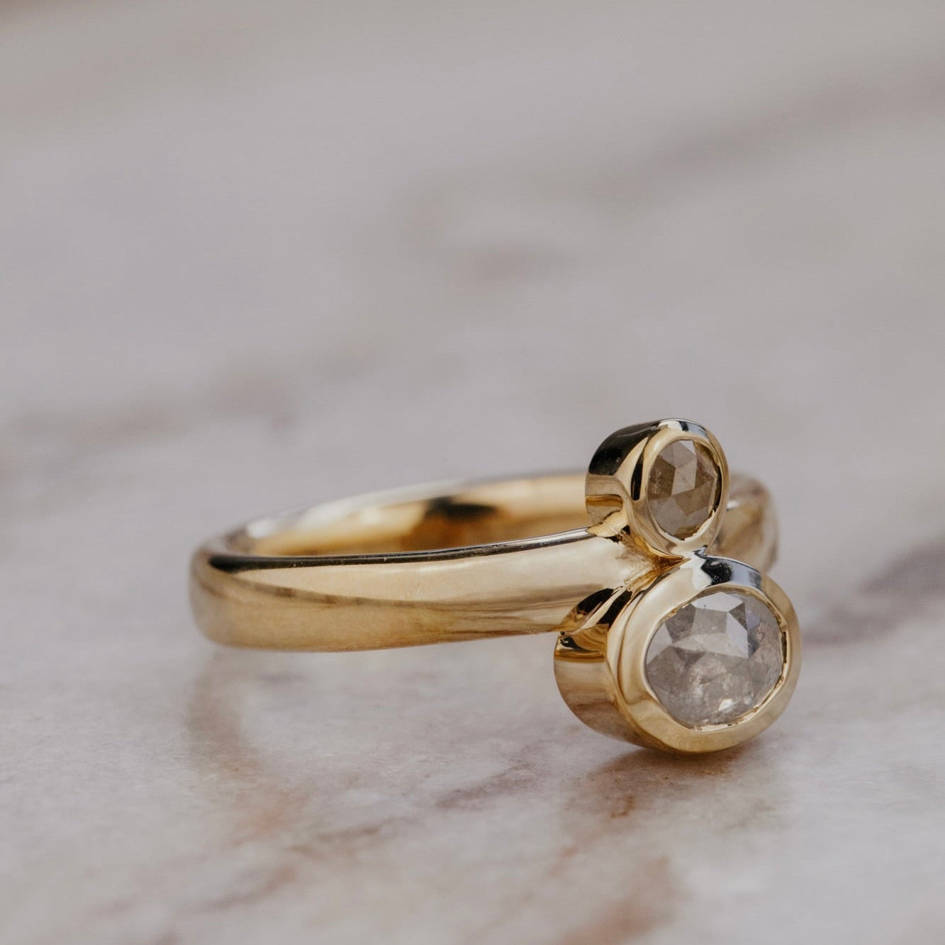 The Reese 1.03 ct Oval and .15 ct Round Rustic Diamond Ring in 14k yellow gold featuring an oval and round rustic diamond set north-south in bezel settings on a wide band, shown at an angle on a marble surface to display the wide band profile and bezel setting depth.