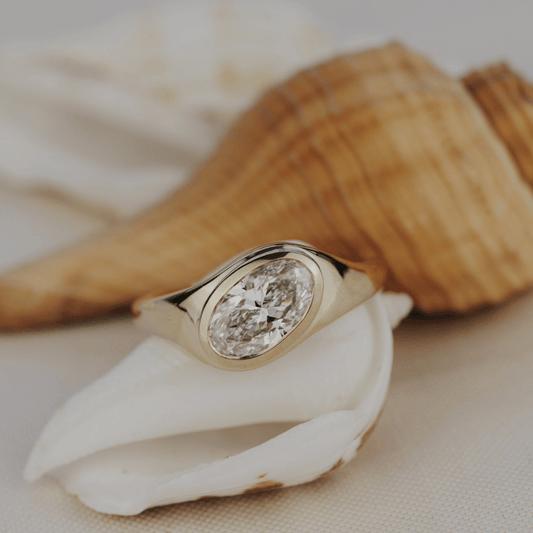 A gold ring with an oval diamond displayed on white seashells with a larger shell blurred in the background.
