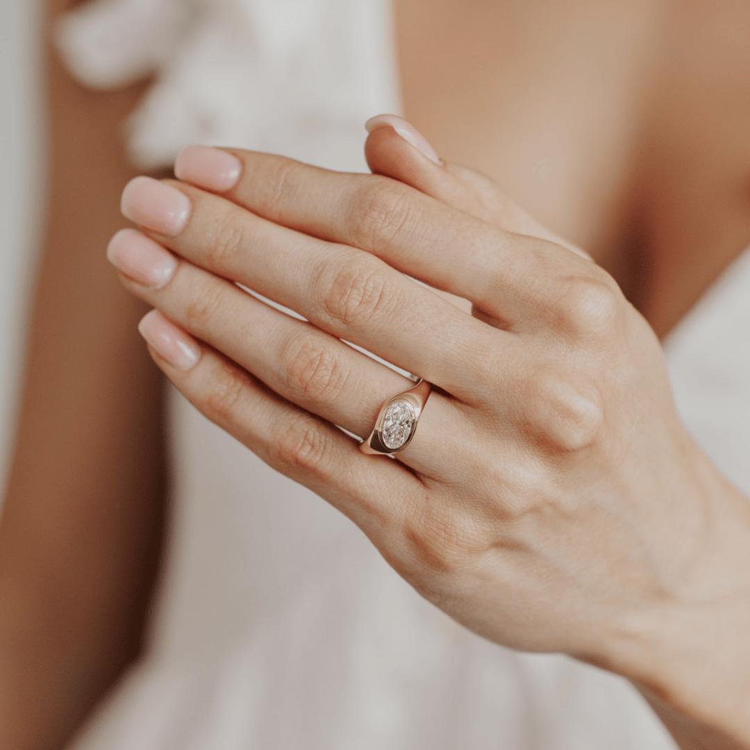 A close-up of a person's hands wearing a delicate yellow gold ring with a oval-cut diamond gemstone. The background is softly blurred, showing a white bridal veil.