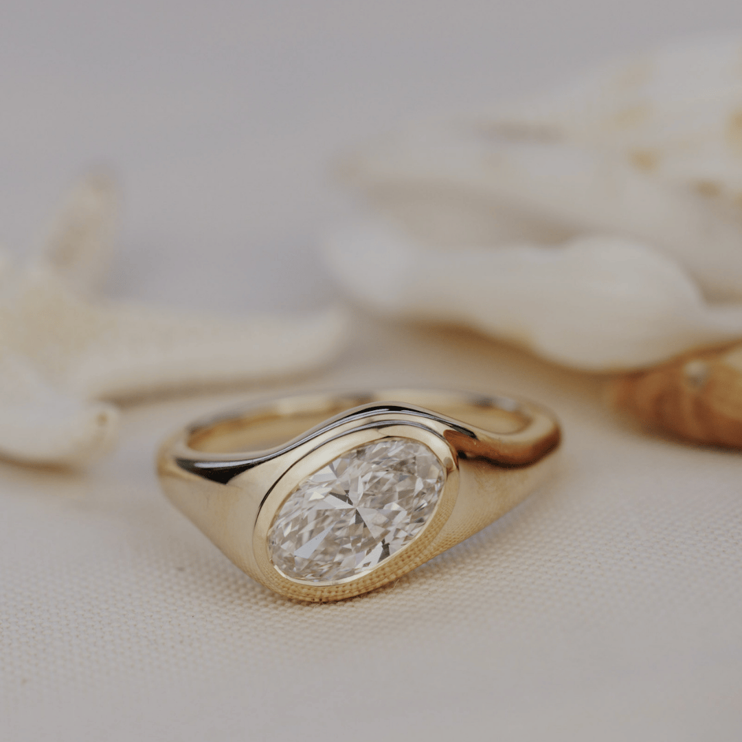 A gold signet ring with an oval-cut diamond, displayed on a textured beige fabric with blurred white seashells in the background.