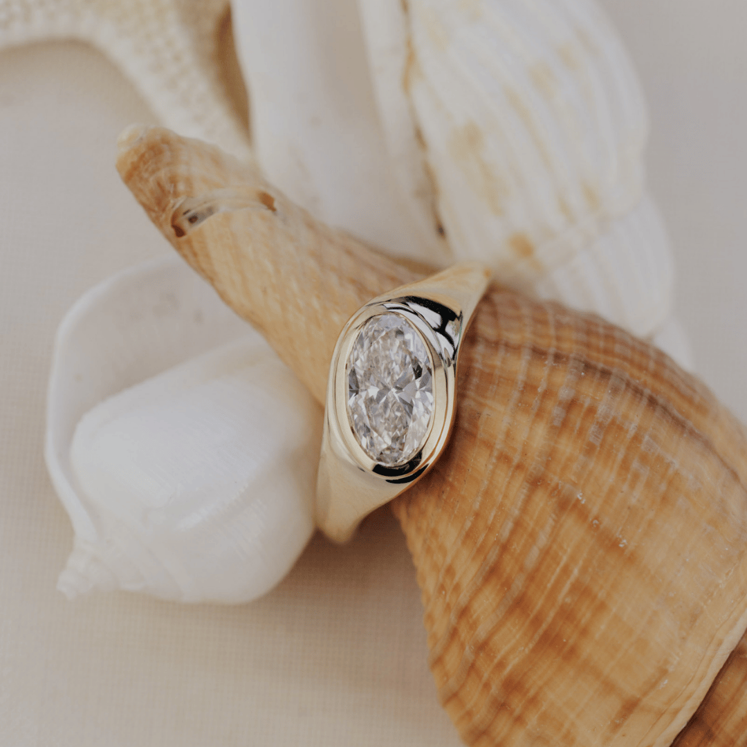 A gold signet ring with an oval-cut diamond, displayed on a large seashell with smaller white shells in the background.