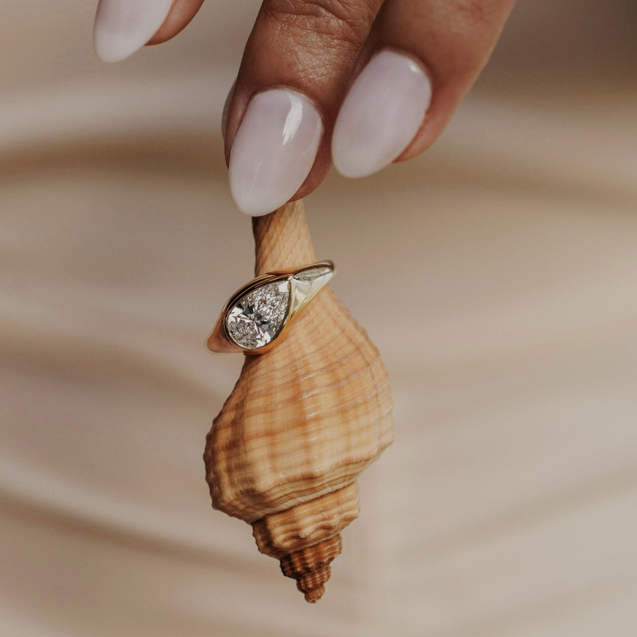 The Santorini Pear Ring in 14k yellow gold featuring a lab-grown pear diamond bezel set on a tapered band, displayed on a seashell held by a model's hand.