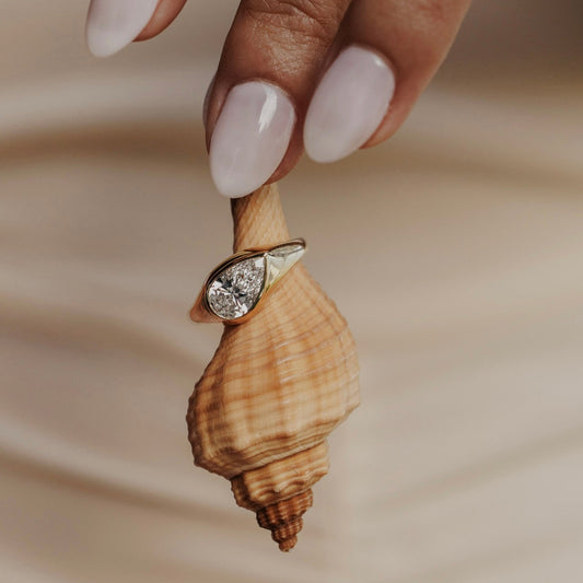 The Santorini Pear Ring in 14k yellow gold featuring a lab-grown pear diamond bezel set on a tapered band, displayed on a seashell held by a model's hand.