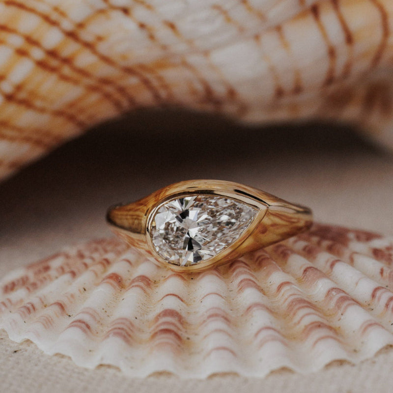 The Santorini Pear Ring in 14k yellow gold featuring a lab-grown pear diamond bezel set on a tapered band, displayed inside a seashell. This 14k gold ring features a pear diamond center stone set in a bezel design on a tapered band. This ring is made-to-order based on the selected customizations.