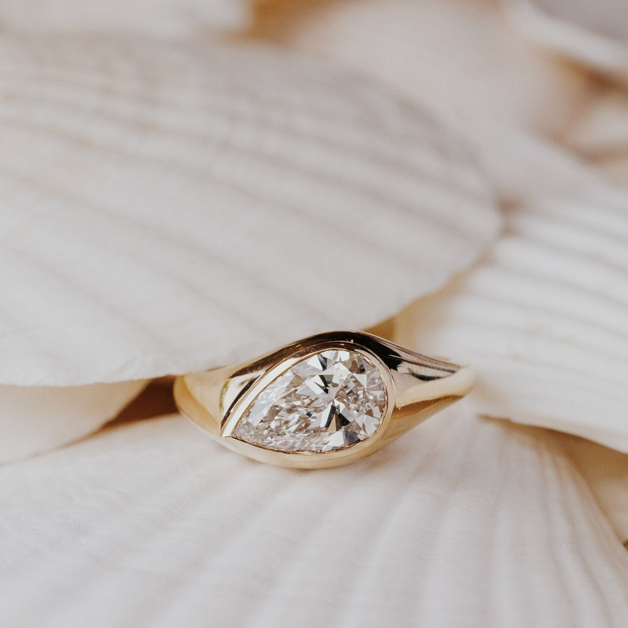 The Santorini Pear Ring in 14k yellow gold featuring a lab-grown pear diamond bezel set on a tapered band, resting on white shells. This 14k gold ring features a pear diamond center stone set in a bezel design on a tapered band. This ring is made-to-order based on the selected customizations.