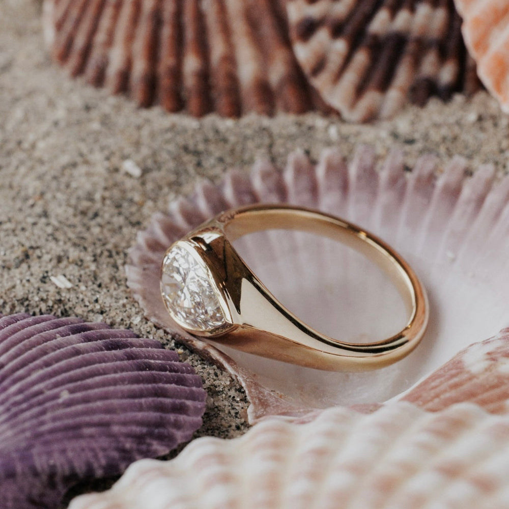The Santorini Pear Ring in 14k yellow gold featuring a lab-grown pear diamond bezel set on a tapered band, displayed among seashells and sand.