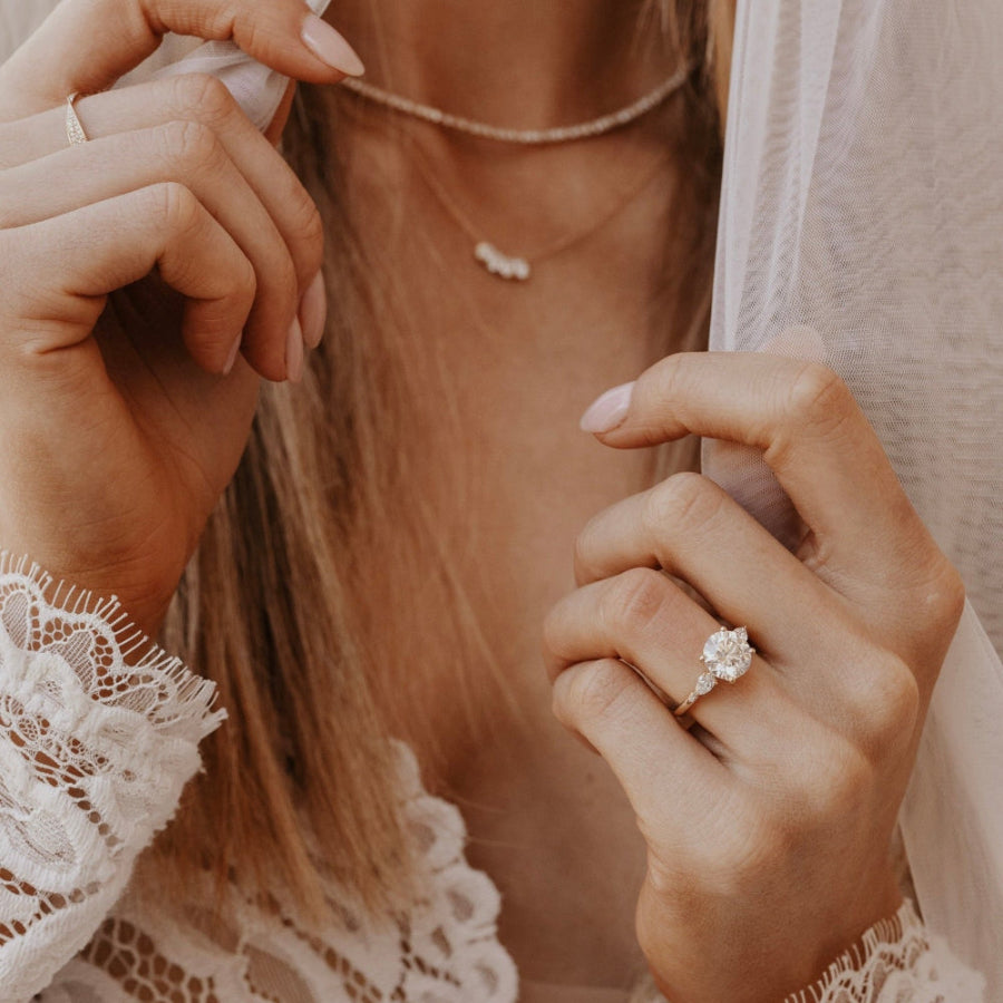 The Toula Round Ring in 14k yellow gold with a lab grown diamond round center stone and pear and round natural diamond side stones, worn on a bride's hand alongside delicate gold necklaces.