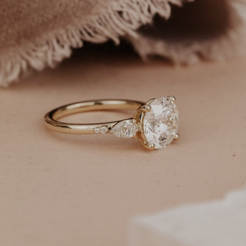 The Toula Round Ring in 14k yellow gold featuring a lab grown diamond round center stone with pear and round natural diamond side stones, shown from a side profile angle on a warm neutral surface with a soft fabric backdrop.