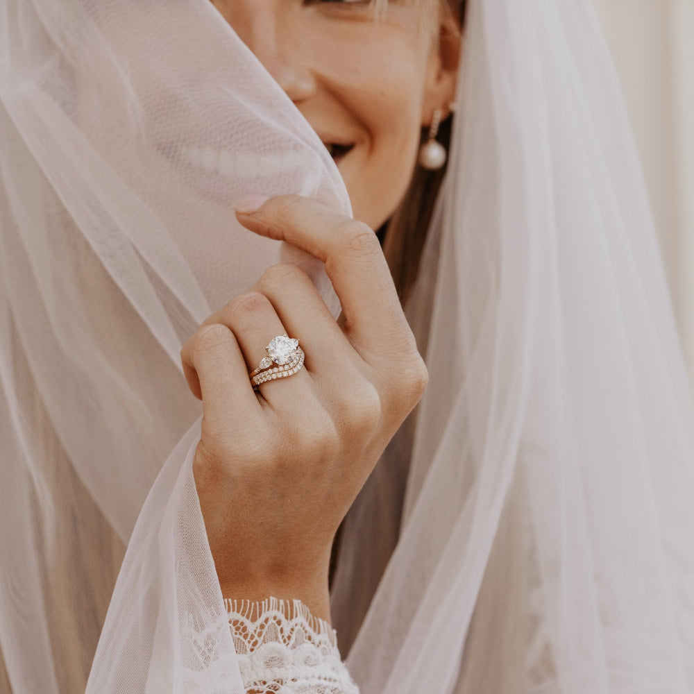 The Toula Round Ring in 14k yellow gold with a lab grown diamond round center stone and pear and round natural diamond side stones, worn stacked with a wedding band on a smiling bride's hand as she holds a white veil.