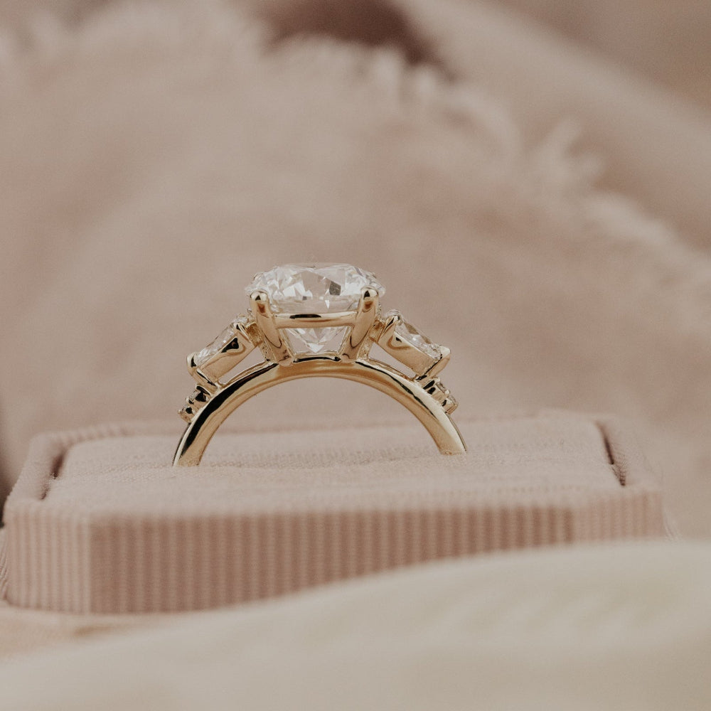 The Toula Round Ring in 14k yellow gold featuring a lab grown diamond round center stone set in four prongs with pear and round natural diamond side stones, displayed facing forward in a blush pink velvet ring box.