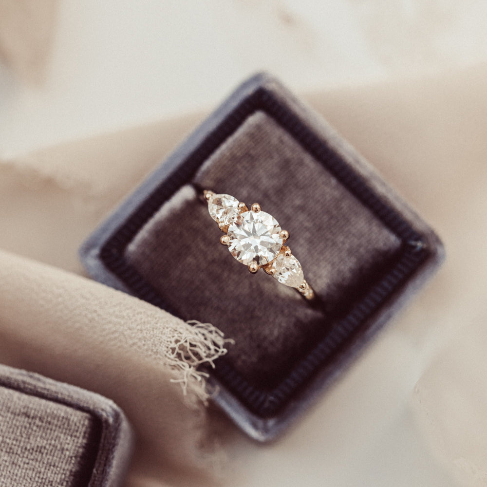 The Toula Round Ring in 14k yellow gold with a lab grown diamond round center stone and pear and round natural diamond side stones, displayed in a charcoal velvet ring box against a soft neutral background.