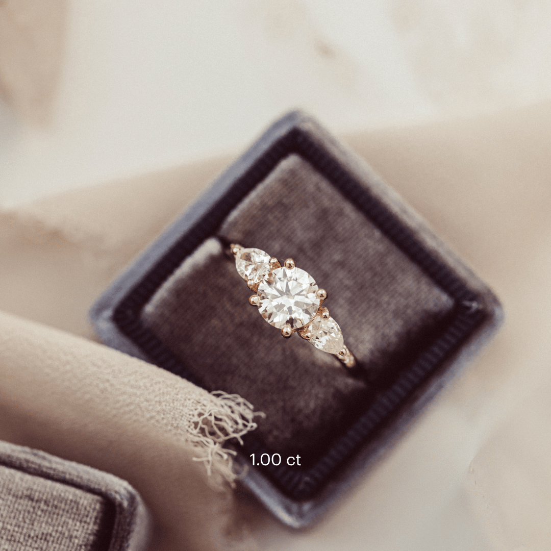 The Toula Round Ring in 14k yellow gold with a lab grown diamond round center stone and pear and round natural diamond side stones, displayed in a charcoal velvet ring box against a soft neutral background.