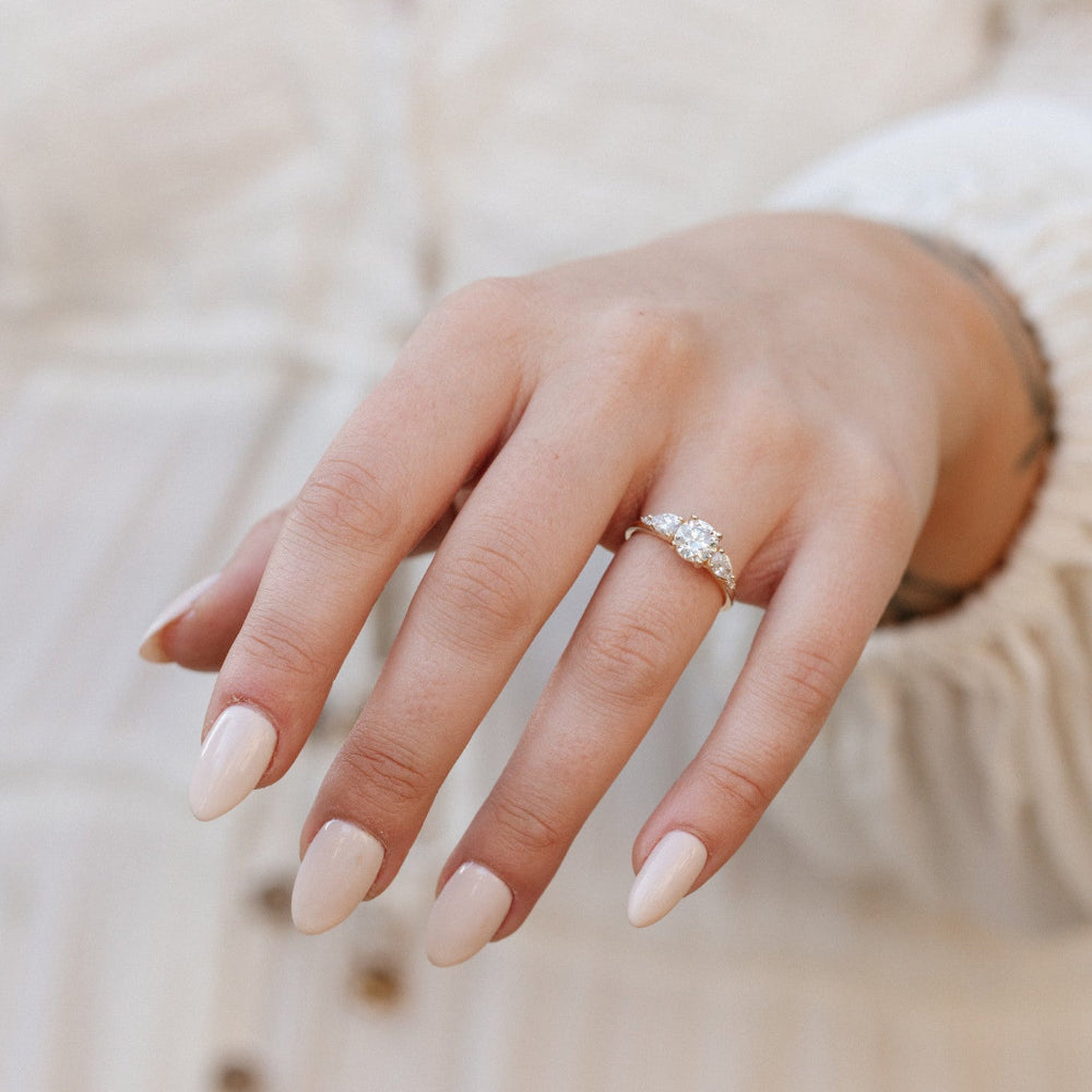 The Toula Round Ring in 14k yellow gold featuring a lab grown diamond round center stone with pear and round natural diamond side stones, worn on a model's outstretched hand against a cream textured background.