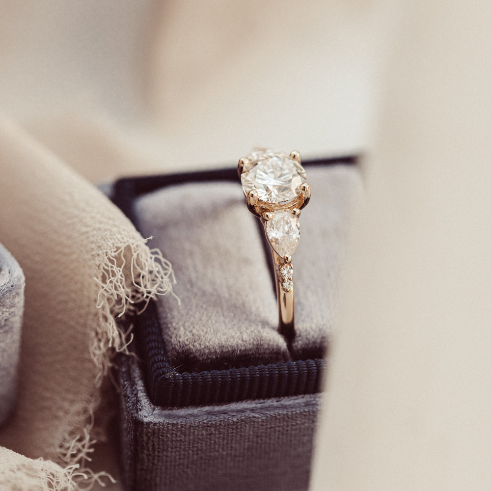The Toula Round Ring in 14k rose gold featuring a lab grown diamond round center stone with pear and round natural diamond side stones, shown from a side angle resting in a charcoal velvet ring box.