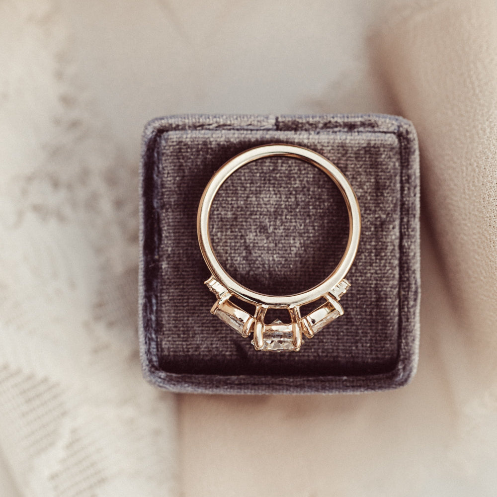 The Toula Round Ring in 14k yellow gold featuring a lab grown diamond round center stone with pear and round natural diamond side stones, displayed from an overhead angle showing the band and stone profile in a charcoal velvet ring box.
