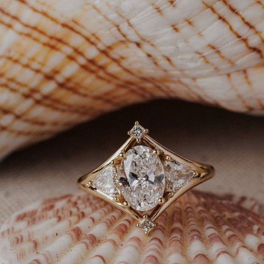 The Tulum Oval Ring in 14k yellow gold featuring a lab-grown oval diamond center stone with trillion and princess cut diamond accents in a geometric halo, displayed inside a seashell. The Tulum Ring is made in 14k gold, featuring an oval center stone, with two trillion diamonds on either side, surrounded by a high polish metal halo and two princess cut diamonds on top and bottom of the center stone. The entire design is set flush to the finger. This ring is made-to-order based on the selected customizations