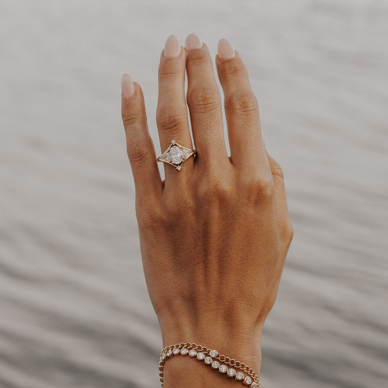 The Tulum Oval Ring in 14k yellow gold featuring a lab-grown oval diamond center stone with trillion and princess cut diamond accents in a geometric halo, worn on the ring finger alongside stacked diamond bracelets.