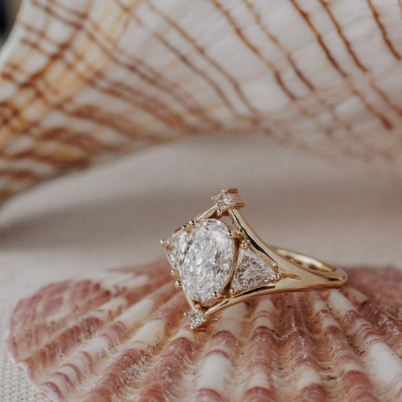 The Tulum Oval Ring in 14k yellow gold featuring a lab-grown oval diamond center stone with trillion and princess cut diamond accents in a geometric halo, displayed inside a seashell. The Tulum Ring is made in 14k gold, featuring an oval center stone, with two trillion diamonds on either side, surrounded by a high polish metal halo and two princess cut diamonds on top and bottom of the center stone. The entire design is set flush to the finger. This ring is made-to-order based on the selected customizations