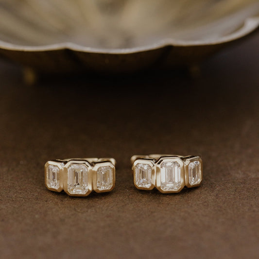 Pair of emerald cut lab grown diamond earrings on a brown surface with a blurred background.
