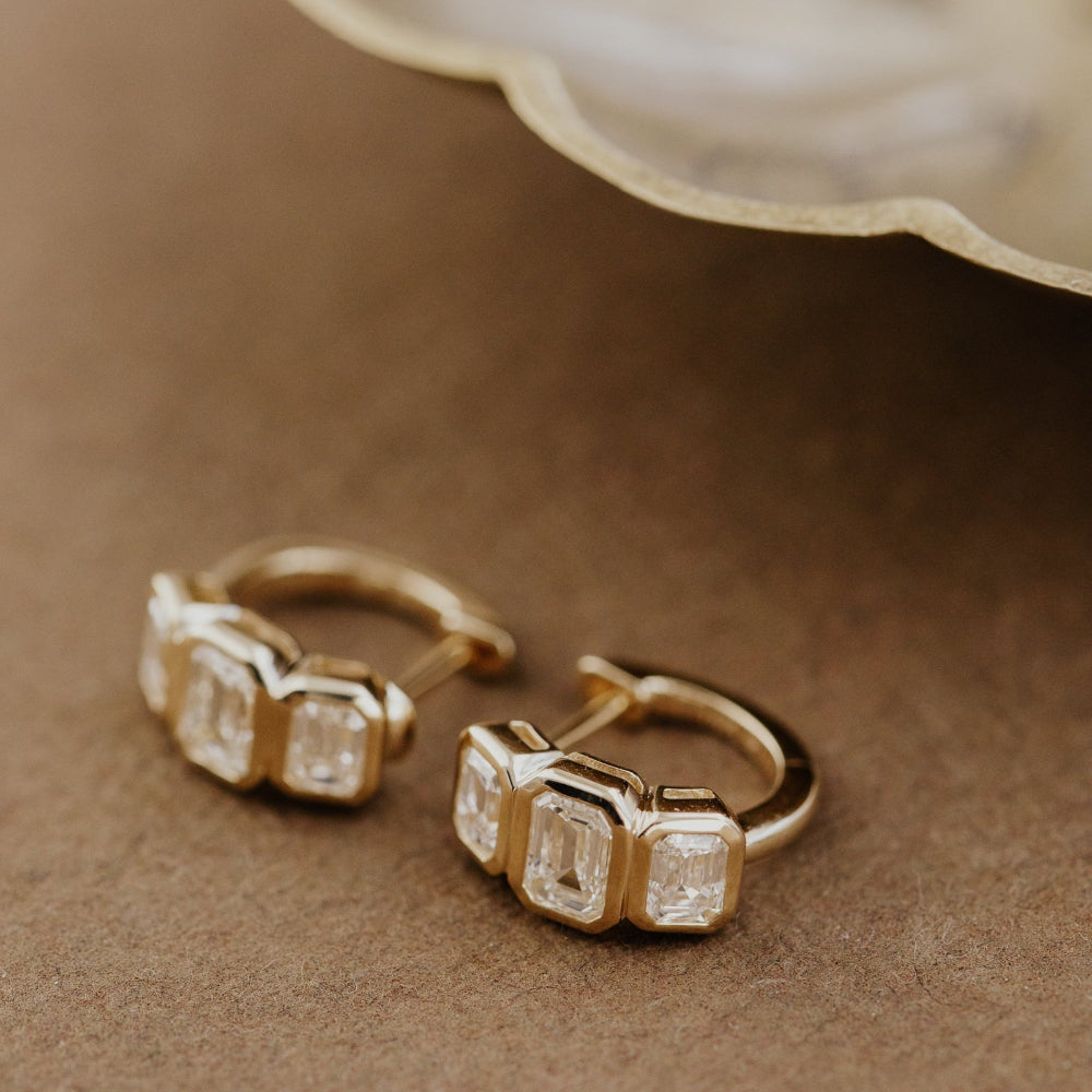 Gold hoop earrings with three emerald cut lab grown diamonds on a brown surface.