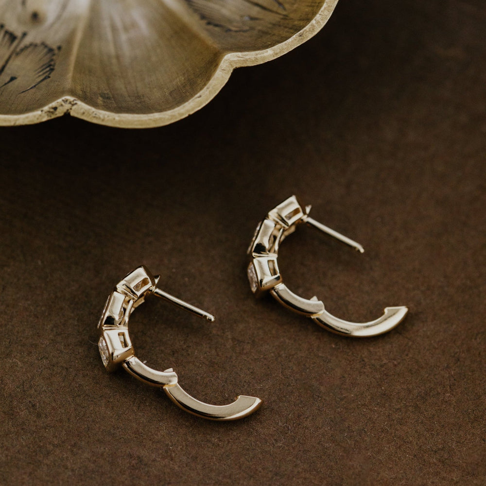 Gold hoop earrings with three lab grown diamonds on a brown surface with a decorative leaf-like object.